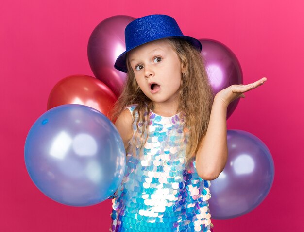 Sorprendido niña rubia con gorro de fiesta azul de pie con globos de helio manteniendo la mano abierta aislada en la pared rosa con espacio de copia