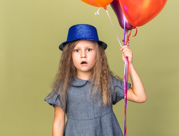 Sorprendido niña rubia con gorro de fiesta azul sosteniendo globos de helio aislado en la pared verde oliva con espacio de copia