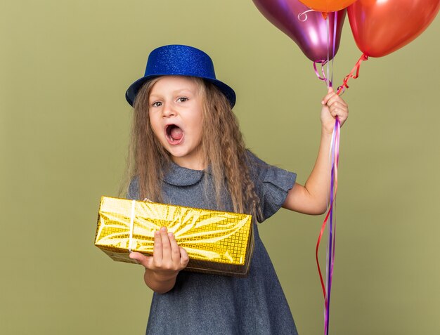 Sorprendido niña rubia con gorro de fiesta azul sosteniendo globos de helio y caja de regalo aislado en la pared verde oliva con espacio de copia