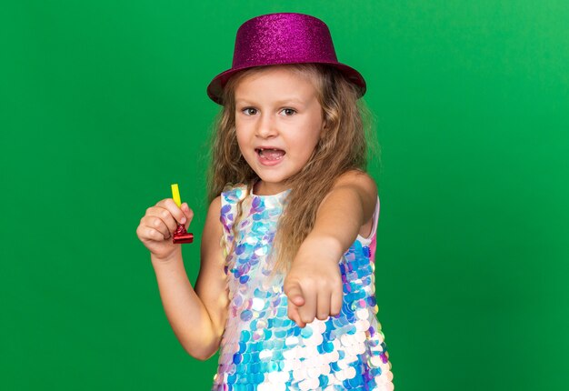 Sorprendido niña rubia con gorro de fiesta púrpura sosteniendo un silbato de fiesta y apuntando aislado en la pared verde con espacio de copia