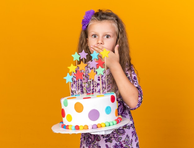 Sorprendido niña rubia sosteniendo la torta de cumpleaños y poniendo la mano en la boca aislada en la pared naranja con espacio de copia