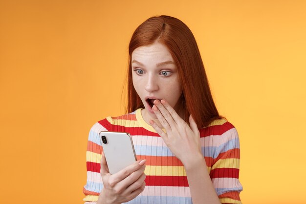 Sorprendido sin palabras joven adolescente pelirroja estudiante jadeando gota mandíbula decir omg wow tapa abierta boca palma mirada sorprendida pantalla del teléfono inteligente leyendo chismes frescos fondo naranja