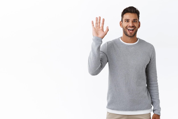 Sorprendido, simpático y extrovertido hombre guapo saludando con la mano saludando sonriendo felizmente, viendo a una persona familiar en cuervos y saludando, vino en la cita del café diciendo hola o hola con un gesto de fondo blanco.