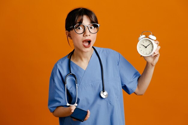 Sorprendido sosteniendo despertador habla por teléfono joven doctora vistiendo uniforme fith estetoscopio aislado sobre fondo naranja