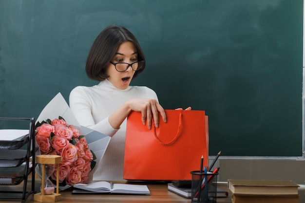 sorprendido sosteniendo y mirando el regalo joven maestra sentada en el escritorio con herramientas escolares en el aula