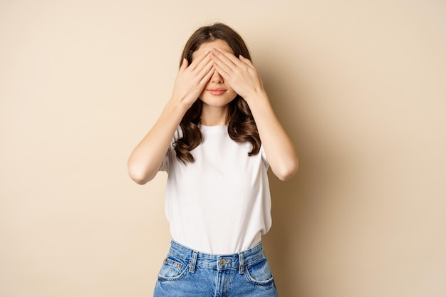 Foto gratuita sorpresa. una joven feliz cerró los ojos con las manos y esperó un regalo, de pie sobre un fondo beige