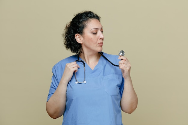 Sospechosa doctora de mediana edad con uniforme y estetoscopio alrededor del cuello agarrando el estetoscopio mirándolo aislado en un fondo verde oliva