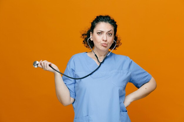 Sospechosa doctora de mediana edad con uniforme y estetoscopio manteniendo la mano en la cintura mirando la cámara agarrando el estetoscopio aislado en el fondo naranja