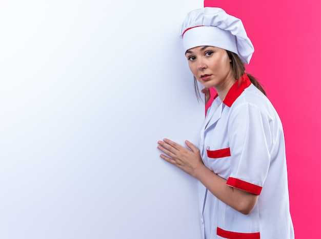 Sospechosa joven cocinera con uniforme de chef se encuentra cerca de la pared blanca aislada sobre fondo rosa con espacio de copia