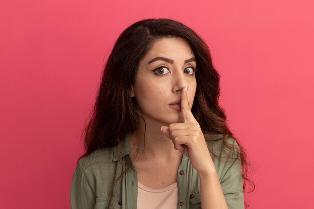 Foto gratuita sospechosa joven hermosa vestida con camiseta verde oliva mostrando gesto de silencio aislado en la pared rosa