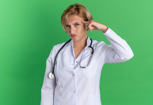 Sospechoso joven doctora vistiendo bata médica con estetoscopio poniendo el dedo en la sien aislado sobre fondo verde