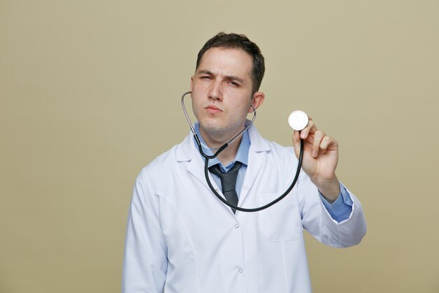 Sospechoso joven médico masculino con bata médica y estetoscopio mirando a la cámara agarrando el estetoscopio mostrándolo aislado en un fondo verde oliva