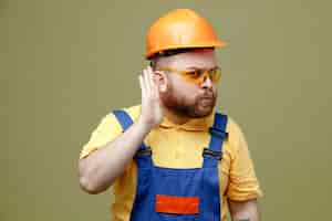 Foto gratuita sospechoso mostrando escuchar gesto joven constructor hombre en uniforme aislado sobre fondo verde