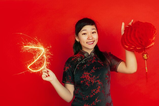 Sosteniendo bengala y linterna. Feliz Año Nuevo Chino. Retrato de joven asiática sobre fondo rojo. Modelo femenino en ropa tradicional se ve feliz. Copyspace.