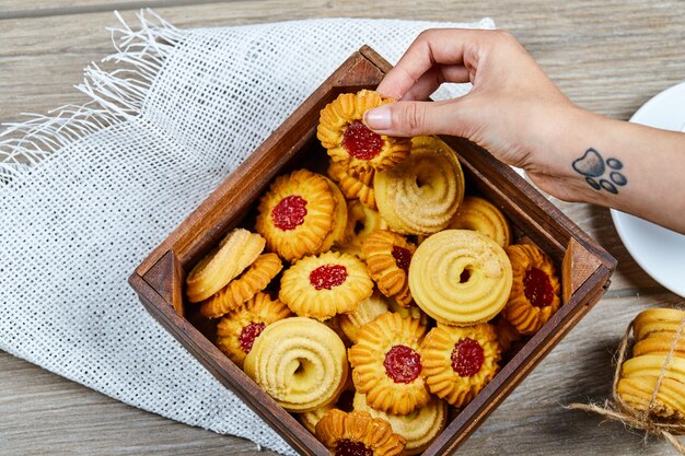 Sosteniendo una galleta y una canasta de galletas variadas sobre la mesa de madera.