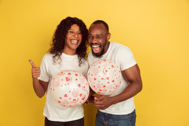 Sosteniendo globos. Celebración del día de San Valentín, feliz pareja afroamericana aislada en la pared amarilla. Concepto de emociones humanas, expresión facial, amor, relaciones, vacaciones románticas.