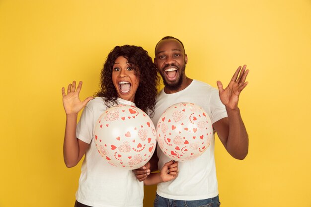 Sosteniendo globos. Celebración del día de San Valentín, feliz pareja afroamericana aislada sobre fondo amarillo de estudio. Concepto de emociones humanas, expresión facial, amor, relaciones, vacaciones románticas.