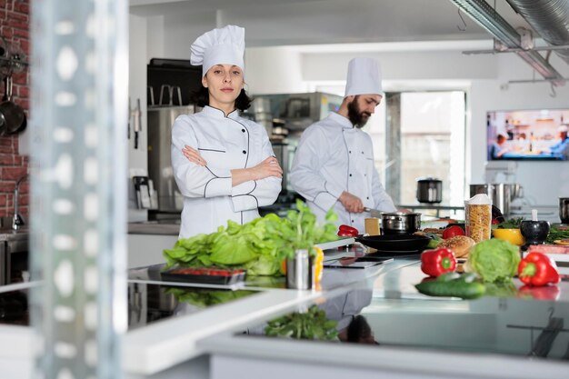 Sous chef confiado parado en el espacio de la cocina profesional del restaurante con los brazos cruzados mientras mira con confianza a la cámara. Jefe de cocina preparando especias e ingredientes para el plato de la cena.