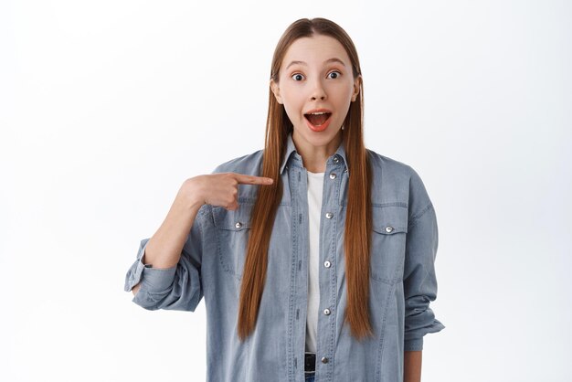 ¿Soy yo? Una chica alegre y sorprendida se señala a sí misma con una cara feliz fascinada al ser elegida ganadora de pie sobre fondo blanco.