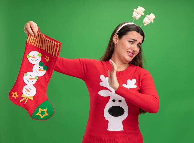 Squeamish hermosa joven vistiendo un suéter de navidad con aro de pelo de navidad sosteniendo calcetines de navidad mostrando gesto de parada aislado sobre fondo verde