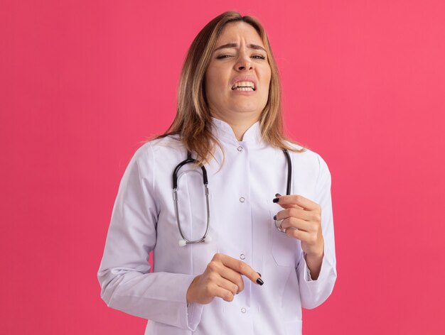 Squeamish joven doctora vistiendo bata médica con estetoscopio aislado en la pared rosa