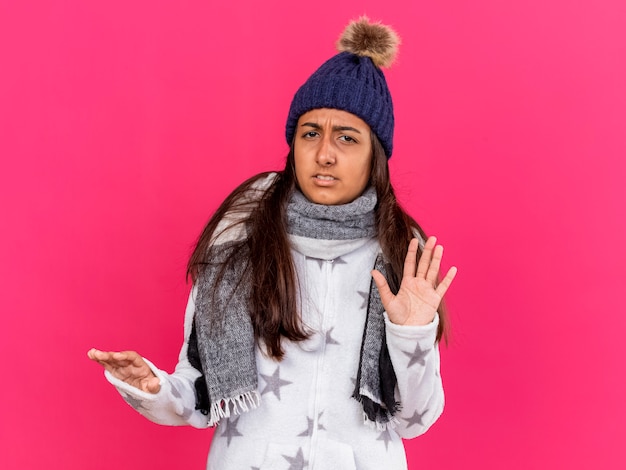 Squeamish joven enferma vistiendo gorro de invierno con bufanda mostrando gesto de parada aislado en rosa