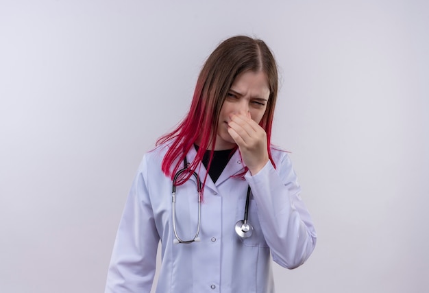 Squeamish joven médico vestida con estetoscopio bata médica nariz cerrada sobre fondo blanco aislado