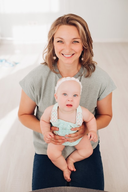 Stock photo retrato de una hermosa madre sonriente en camiseta gris y jeans sosteniendo a su pequeña y encantadora hija en sus manos y mirando a la cámara felizmente ....