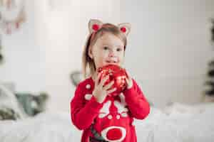 Foto gratuita stock photo retrato de niña adorable en vestido rojo con estampado festivo sosteniendo un regalo dorado bellamente envuelto en las manos mientras está sentado en el suelo junto al árbol de navidad decorado con guirnalda.