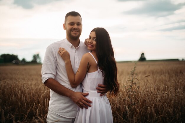 Stock photo retrato de novio barbudo abrazando a su hermosa novia ambos en ropa blanca abrazándose en campo de trigo. Hermoso campo de trigo en el fondo.