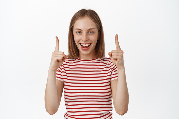 Su producto a continuación. Mujer joven feliz riendo, sonriendo y señalando con el dedo hacia arriba, mostrando pancartas de venta, publicidad debajo de su cabeza, parada en una camiseta de verano contra fondo blanco.