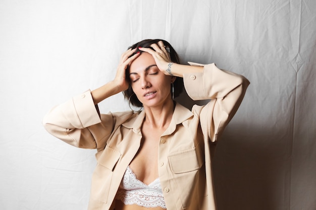 Suave retrato de una bella mujer joven con grandes pechos en un traje beige y sujetador de encaje blanco se sienta en una silla en un blanco gris