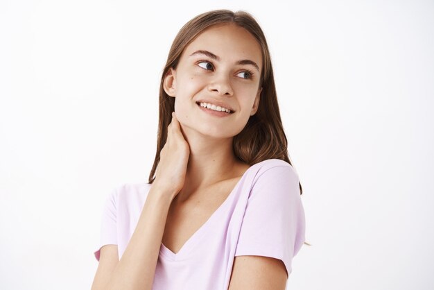 Suave y tierna morena femenina joven en camiseta casual tocando el cuello y mirando a la derecha con una linda sonrisa sensual que se siente bonita con la piel limpia y pura después de aplicar la crema diaria