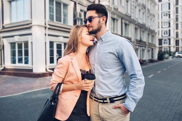 Suavemente la pareja se abraza en la calle de la ciudad. Un chico guapo es camisa azul y gafas de sol se ve seriamente, una chica rubia bonita con un vestido negro abrazándolo.