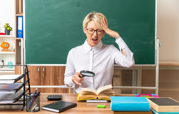 Subrayó joven rubia profesora con gafas sentado en el escritorio con herramientas escolares en el aula sosteniendo lupa sobre libro abierto manteniendo la mano en la cabeza gritando con los ojos cerrados