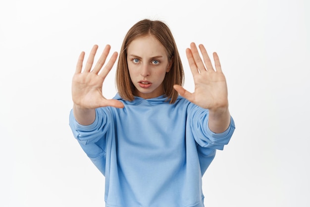 Suficiente, detente ahora. Una joven seria, una adolescente extiende las manos para prohibir, rechazar una mala oferta, mirar con desaprobación, pararse en un fondo blanco y prohibir algo.