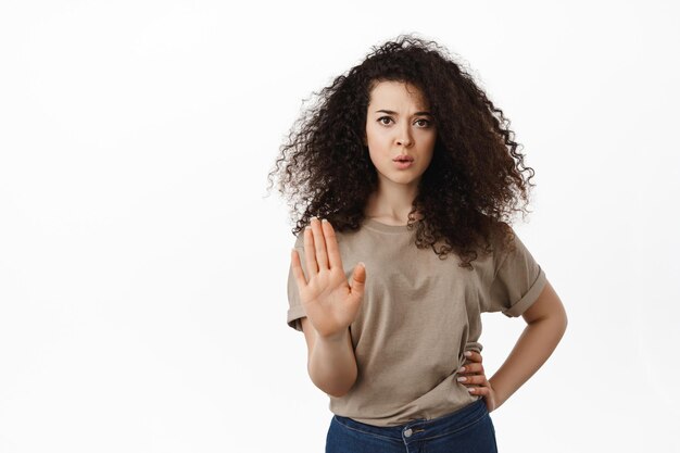 Suficiente, dije que no. Una joven seria detiene a la persona, muestra un gesto de desaprobación en bloque, extiende la palma para rechazar la oferta, se mantiene alejada, el concepto de distanciamiento social, de pie sobre fondo blanco