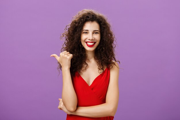 Sugiero que compruebes. Encantadora mujer europea alegre y amistosa con estilo con peinado rizado en vestido rojo de noche apuntando a la izquierda con el pulgar y sonriendo seguro y confiado mostrando copia espacio.