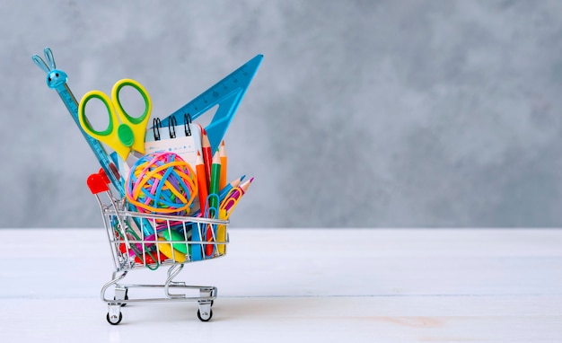 Suministros escolares multicolores en un carrito de compras sobre un fondo gris con espacio de copia de texto. El concepto de regresar a la escuela para el nuevo año escolar, Compras.