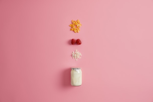 Superalimento saludable y concepto de nutrición orgánica. Tarro de yogur fresco y tres ingredientes nutritivos para preparar el desayuno. Copos de granola, frambuesa roja y coco sobre fondo rosa.