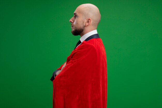 Superhéroe barbudo hombre de negocios en capa roja de pie de lado con cara seria sobre pared verde