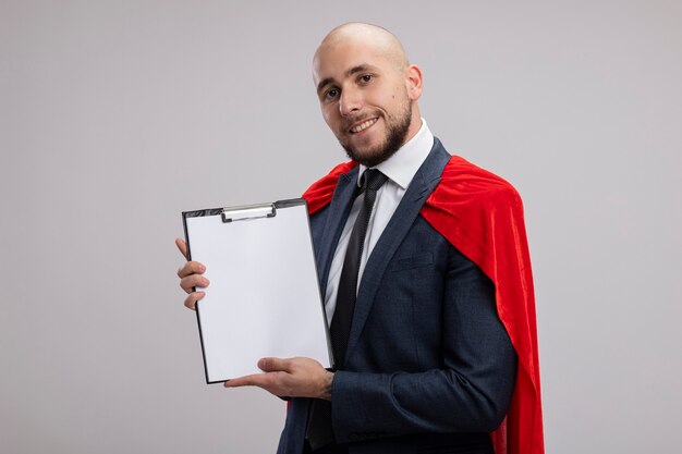 Superhéroe barbudo hombre de negocios en capa roja que muestra el portapapeles con páginas en blanco con una sonrisa en la cara de pie sobre la pared blanca