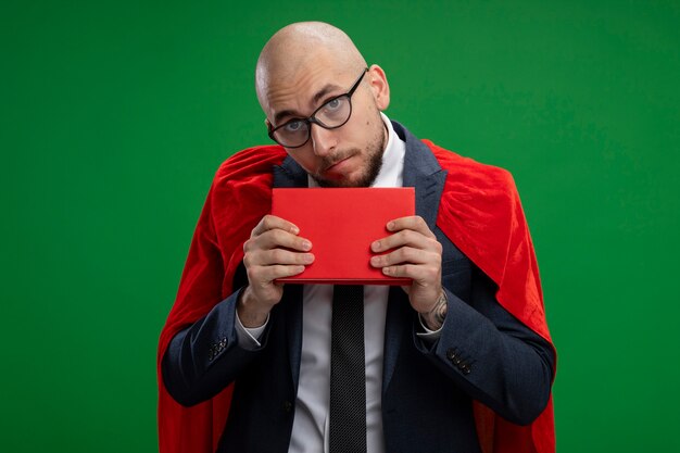 Superhéroe barbudo hombre de negocios en capa roja sosteniendo libro confundido y muy ansioso de pie sobre la pared verde