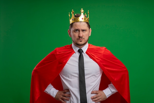 Foto gratuita superhéroe empresario en capa roja con corona con rostro serio con los brazos en la cadera de pie sobre la pared verde