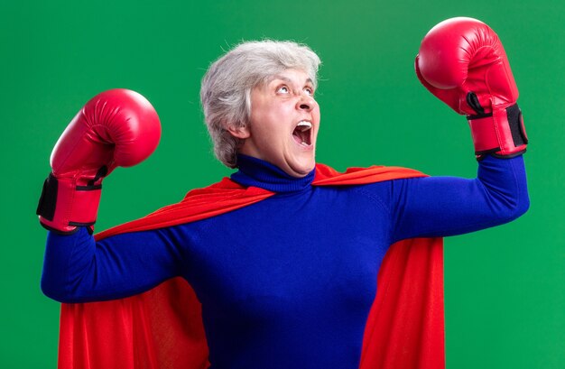 Superhéroe mujer Senior vistiendo capa roja con guantes de boxeo posando como un ganador emocionado