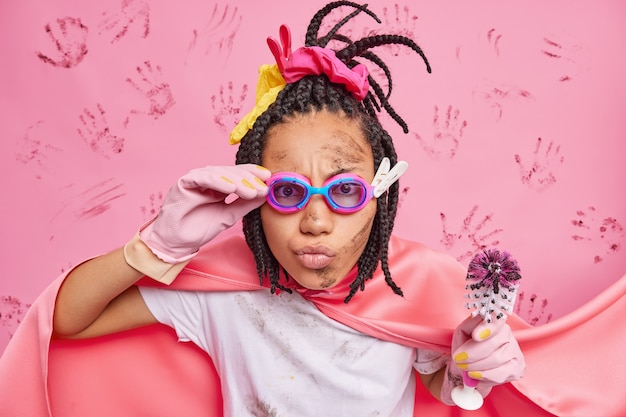 Supermujer escrupulosa lista para salvar el mundo de la suciedad limpia el inodoro con un cepillo mira atentamente a través de gafas tiene la cara sucia vestida con un traje de superhéroe aislado sobre una pared rosa