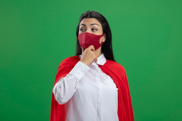 Foto gratuita supermujer joven pensativa con máscara tocando la barbilla mirando al lado aislado en la pared verde