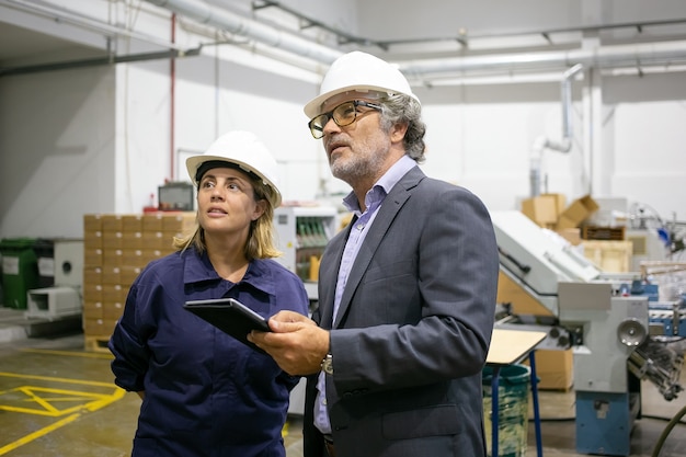 Supervisor barbudo seguro hablando con trabajadora de la planta y sosteniendo la tableta