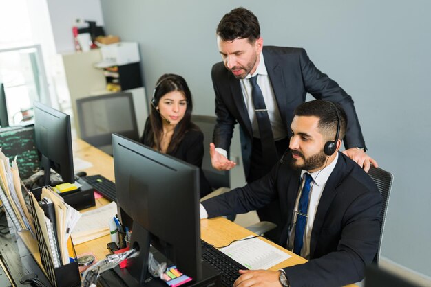 Supervisor masculino entrenando a un ejecutivo latino en un centro de llamadas. Gerente explicando cosas de trabajo a los empleados que ofrecen soporte técnico y servicio al cliente