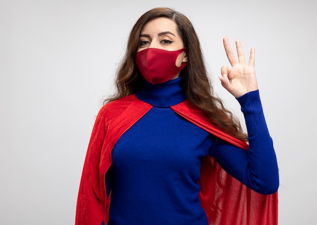 Superwoman complacida con capa roja con máscara protectora roja gestos signo de mano ok aislado en la pared blanca